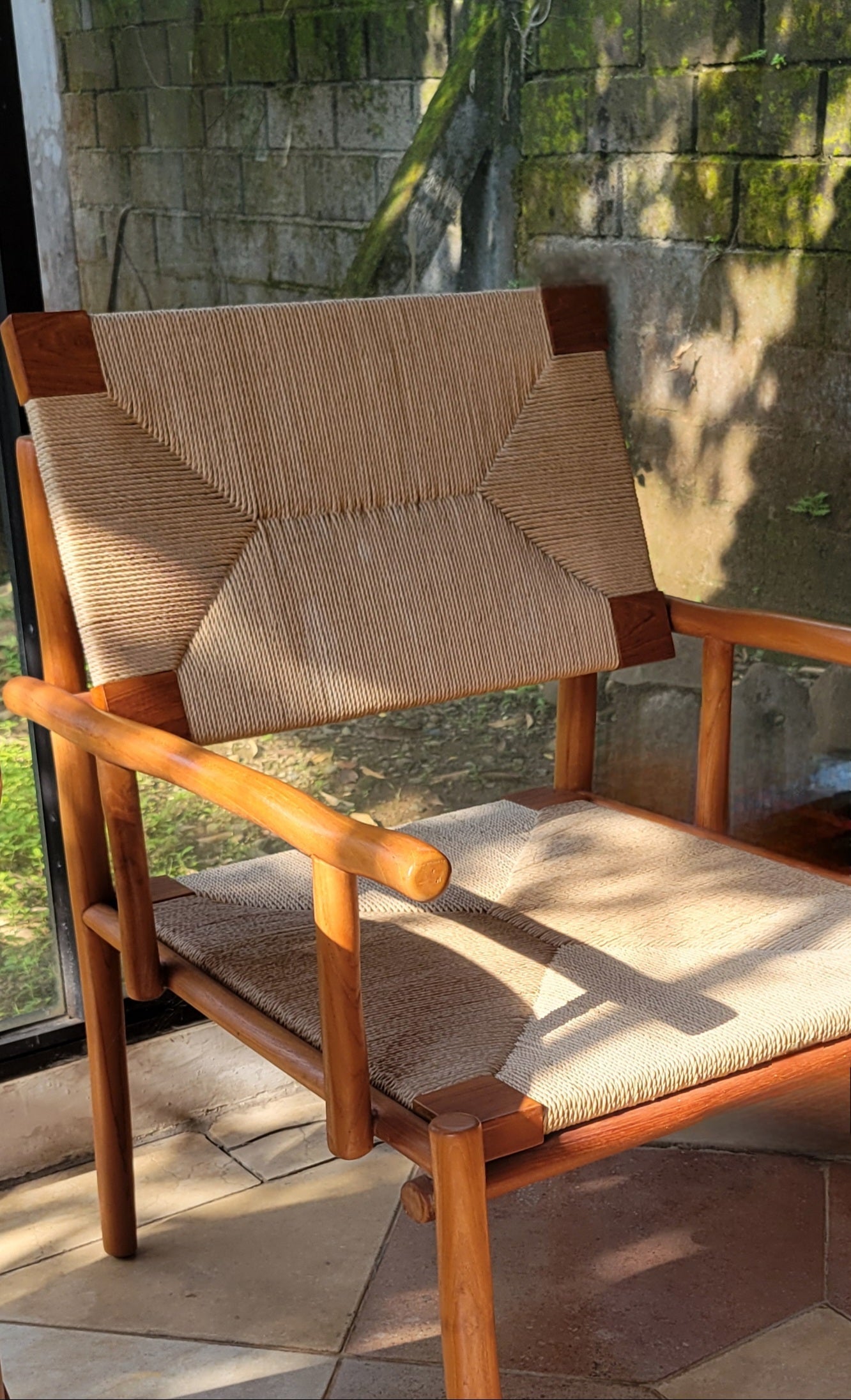 A handcrafted teak chair with a warm, natural wood frame and woven rope seat and backrest. The geometric weave pattern adds texture, while sunlight highlights the smooth teak finish in a terrace setting.