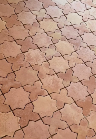 A close-up view of handmade terracotta floor tiles arranged in a repeating geometric star pattern. The tiles feature warm earthy tones with natural color variations and a slightly textured, rustic surface.