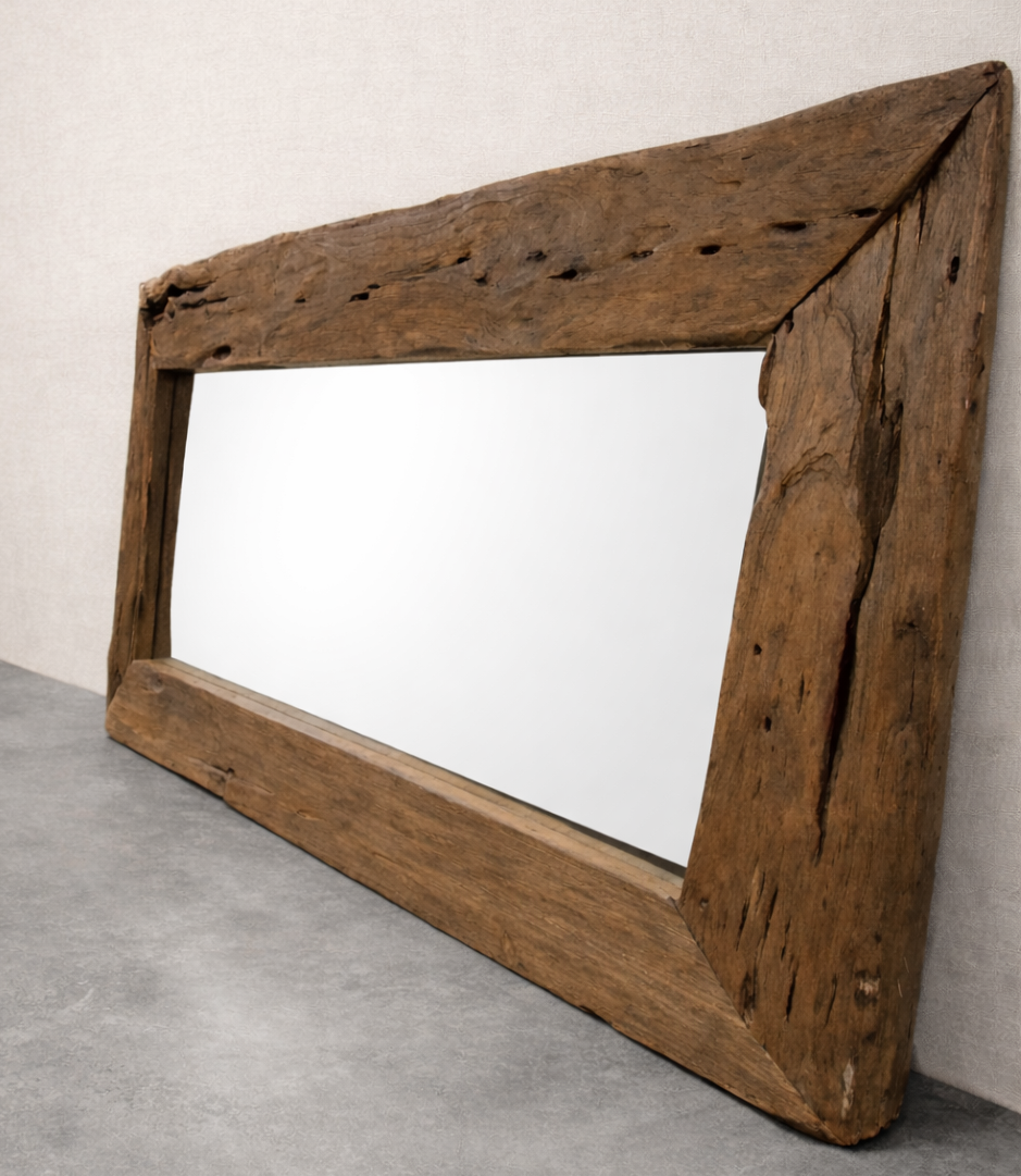 A rustic, rectangular driftwood mirror leans against a soft linen-colored wall on a smooth concrete floor. The weathered wooden frame shows natural cracks and knots, and the mirror reflects a clean, blank surface.