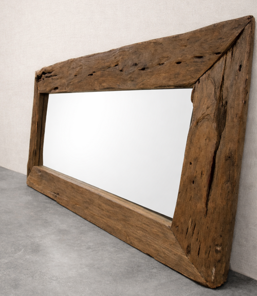 A rustic, rectangular driftwood mirror leans against a soft linen-colored wall on a smooth concrete floor. The weathered wooden frame shows natural cracks and knots, and the mirror reflects a clean, blank surface.