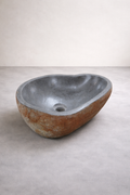 Stone vessel sink with a rough reddish-brown exterior and smooth gray interior in a minimalist studio with a light concrete floor and linen-colored wall.