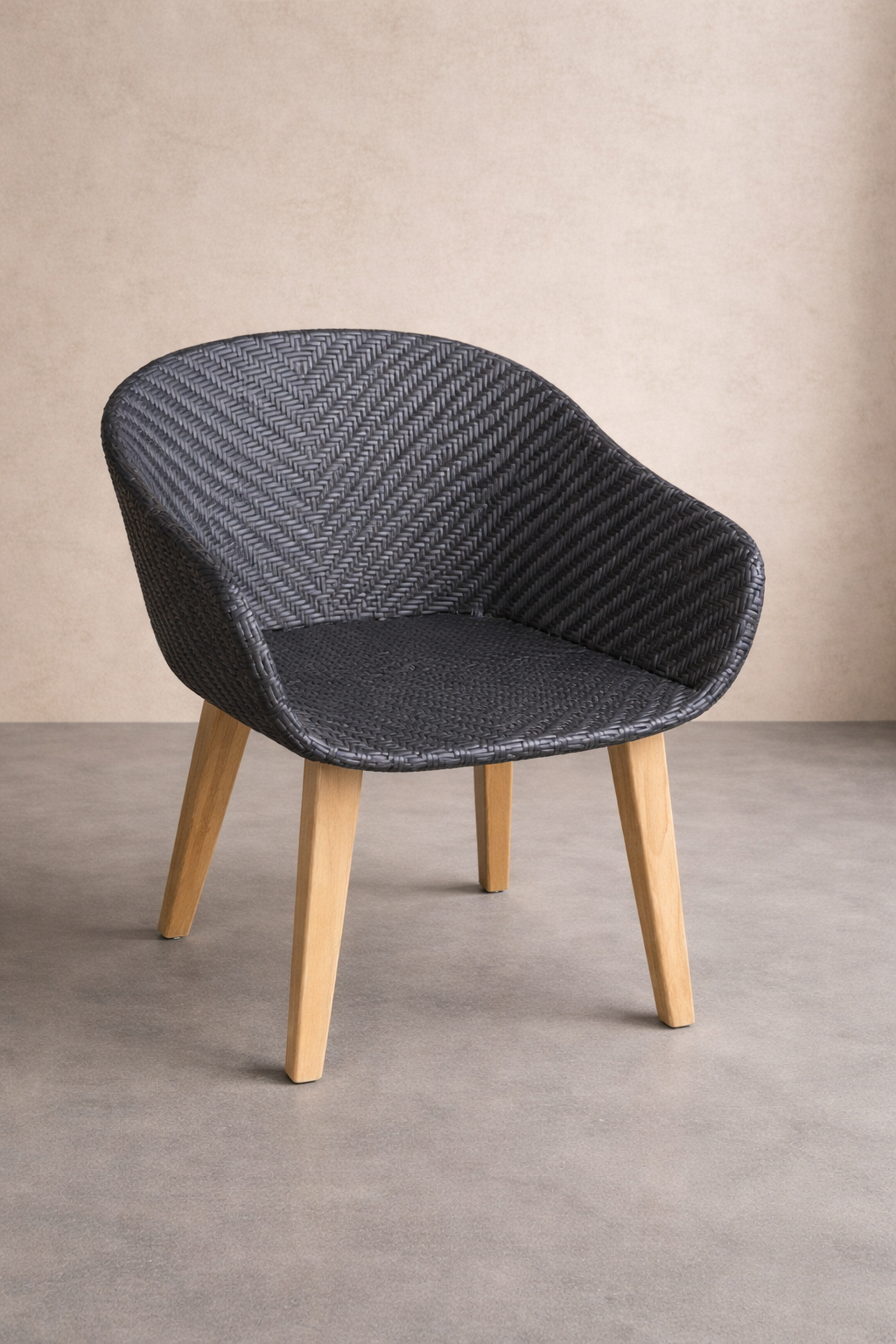 A dark woven synthetic rattan chair with a curved back and seat sits on light wooden legs, placed on a smooth grey concrete floor against a textured linen-colored wall, softly lit in a calm, minimalist studio setting.
