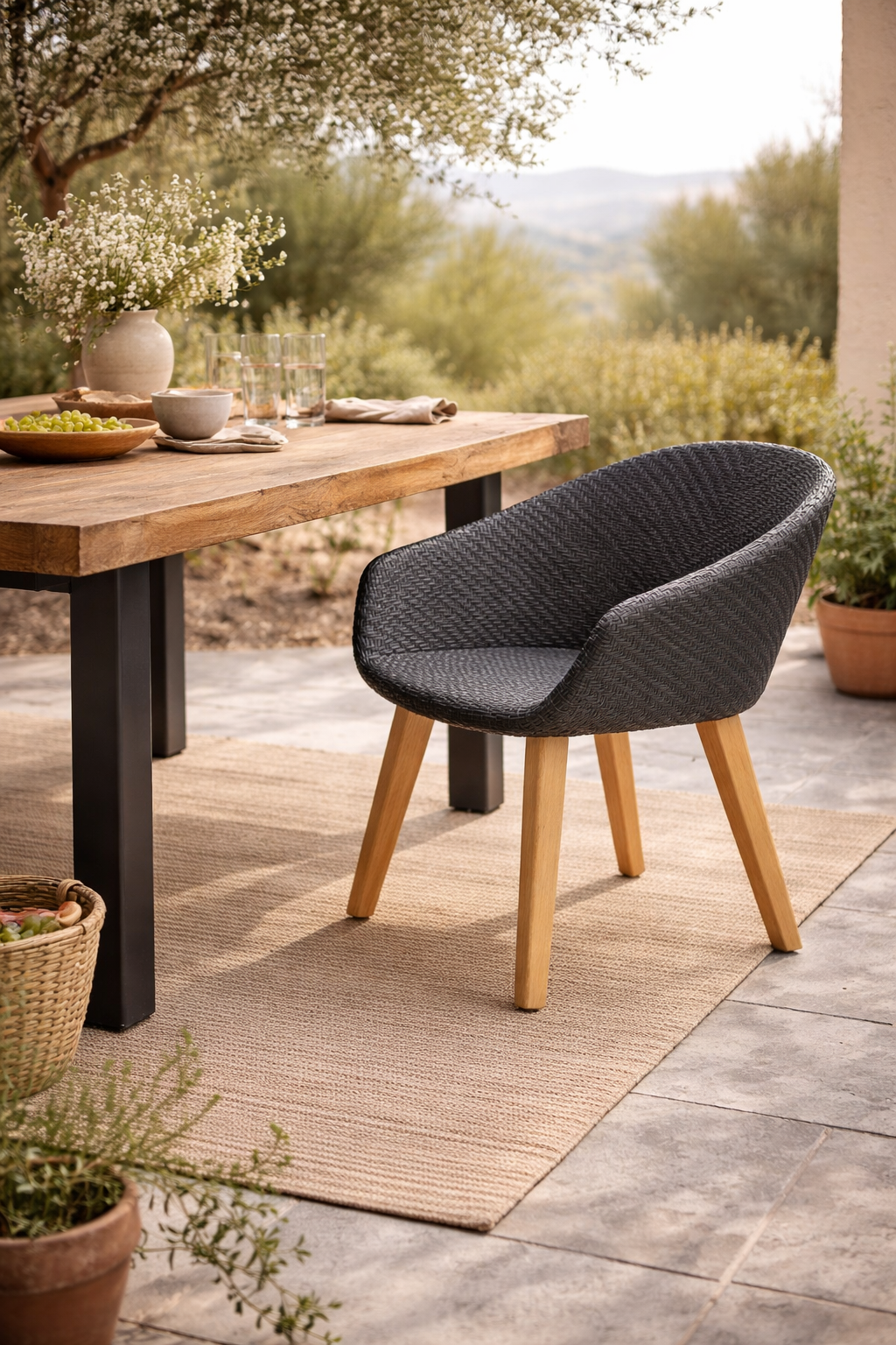 A dark woven rattan chair with light wooden legs is angled toward a reclaimed teak table with black metal legs, set on a natural rug outdoors. Soft sunlight, greenery, and simple table settings create a warm, calm atmosphere.