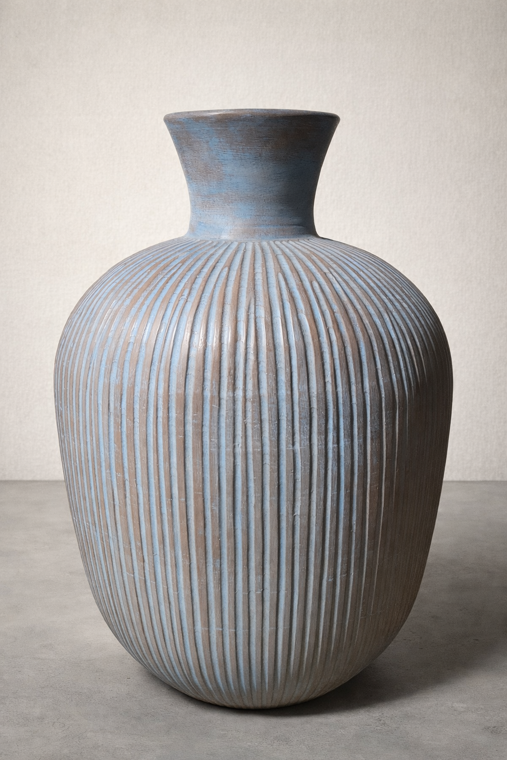 Large ribbed terracotta vase in muted blue and earthy brown tones, photographed in a studio setting on a smooth concrete floor against a soft linen-colored wall, with gentle lighting highlighting its texture.