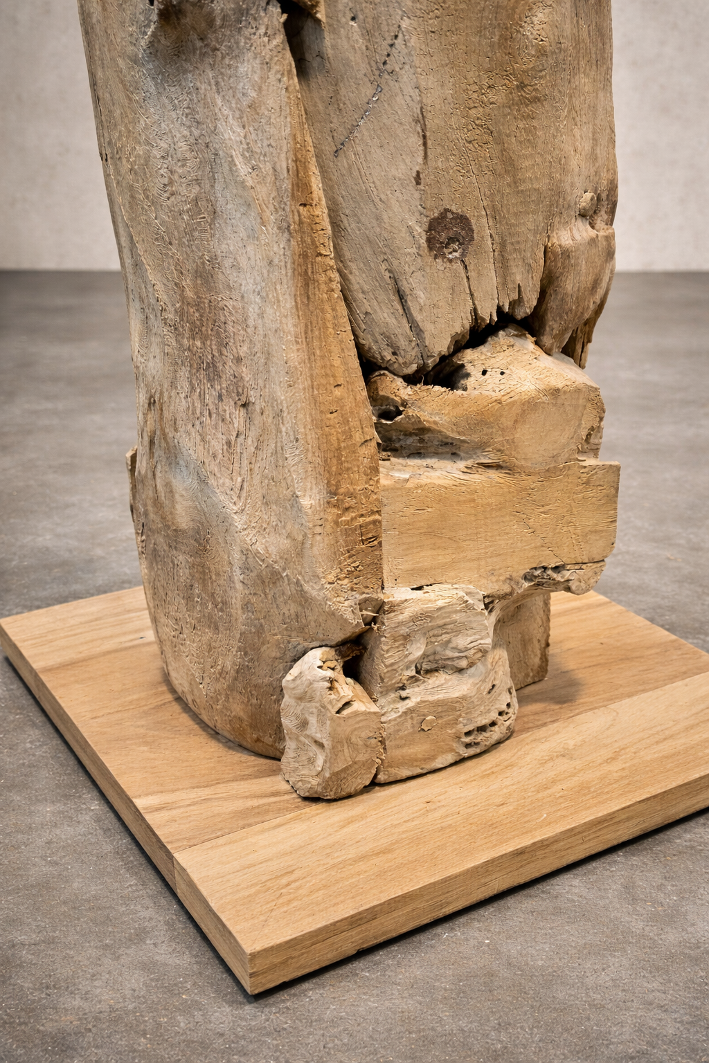 Close-up of the base of a rustic driftwood lamp, showing layered, weathered wood resting on a square wooden platform. Natural textures and warm tones stand out against a linen-colored wall and smooth concrete studio floor.