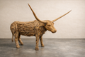 Life-size longhorn sculpture made from reclaimed teak wood, shown in a studio on a concrete floor. Natural layered wood texture, wide curved horns.