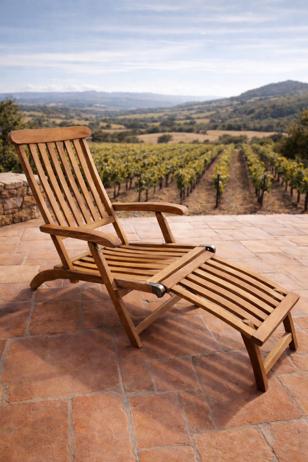 A Teak reclining lounge chair with a footrest sits on warm terracotta tiles, overlooking rows of vineyard vines and rolling hills. Soft sunlight highlights the natural wood, creating a calm, open-air setting with a distant countryside view.
