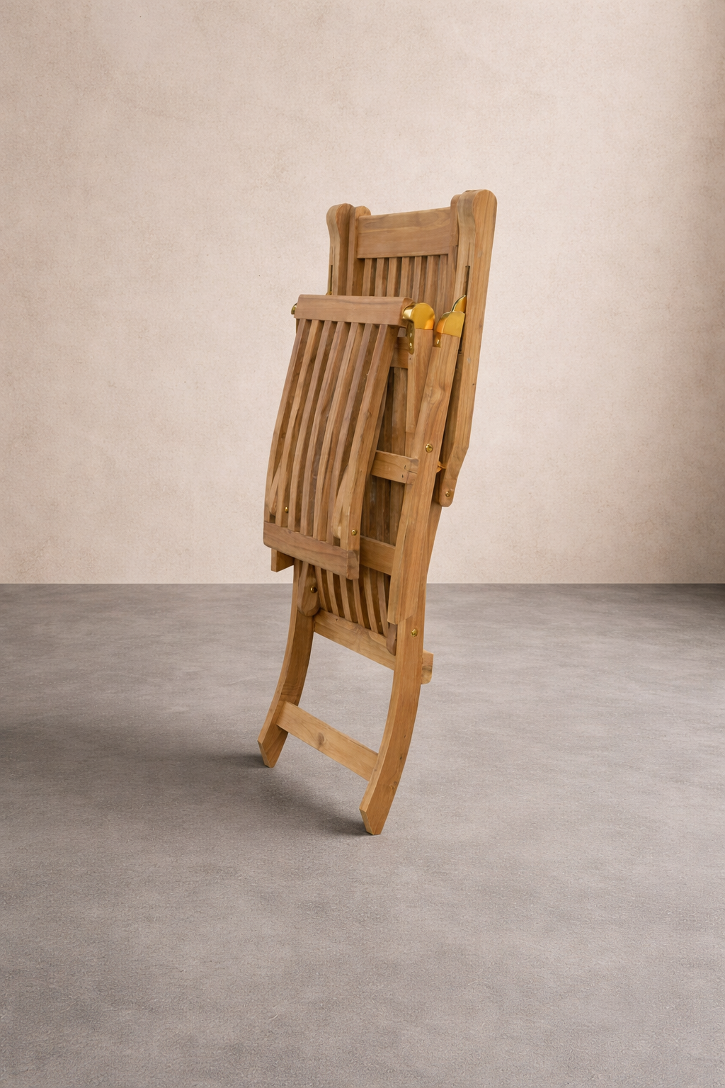 A folded teak lounge chair stands upright on a smooth grey concrete floor against a textured linen-colored wall. Soft studio lighting highlights the slatted wood design and warm tones in a clean, minimalist setting.