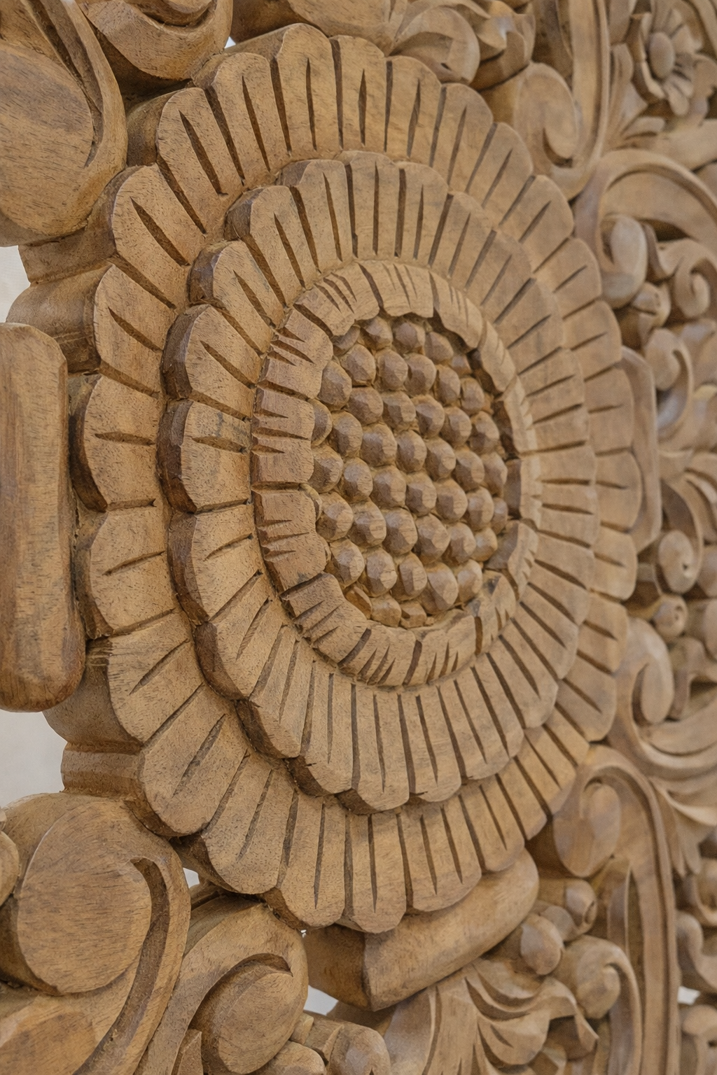 Close-up of an intricately hand-carved wooden mandala panel, showing layered floral petals and textured details in warm teak tones, photographed in soft studio light against a neutral linen-colored background.
