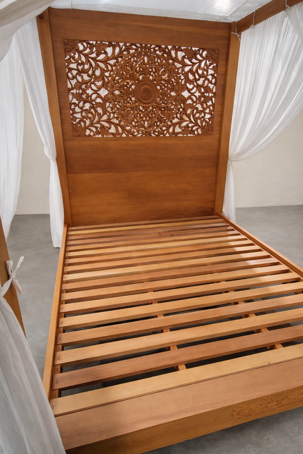 Hand-carved mahogany canopy bed with slatted base and ornate mandala headboard, shown with sheer white drapery and warm natural wood tones in a minimalist studio setting.