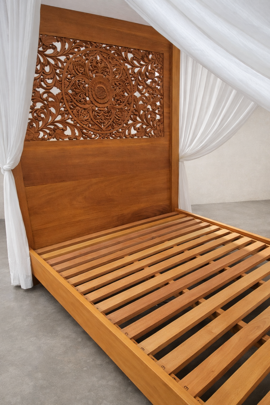 Hand-carved Mahogany canopy bed with slatted base and ornate mandala headboard, framed by sheer white curtains, set on a smooth concrete floor against a soft linen-colored studio wall.