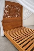 Hand-carved Mahogany canopy bed with slatted base and ornate mandala headboard, framed by sheer white curtains, set on a smooth concrete floor against a soft linen-colored studio wall.