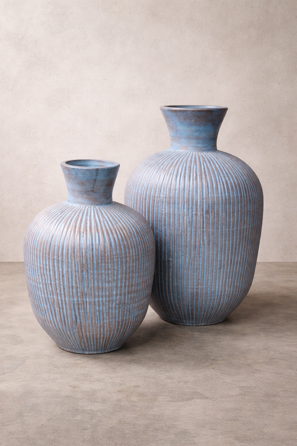 Two tall ribbed ceramic vases in muted blue tones stand side by side on a smooth concrete floor against a linen-colored studio wall, softly lit to highlight their textured vertical grooves and rounded forms.