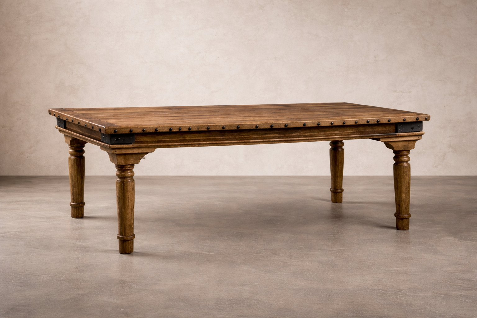 A large rustic wooden table with carved legs and metal stud accents stands on a smooth concrete floor against a textured linen-colored wall, softly lit to highlight its rich grain and handcrafted details.