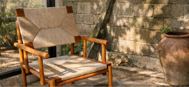 A handcrafted teak chair with a warm, natural wood frame and woven rope seat and backrest. The geometric weave pattern adds texture, while sunlight highlights the smooth teak finish in a terrace setting.