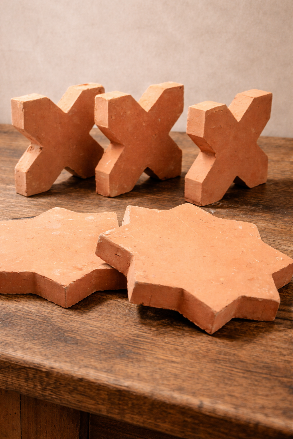 Terracotta star and cross tiles displayed on an aged wooden table, with three cross tiles standing upright behind two star tiles, against a soft linen-colored studio wall.