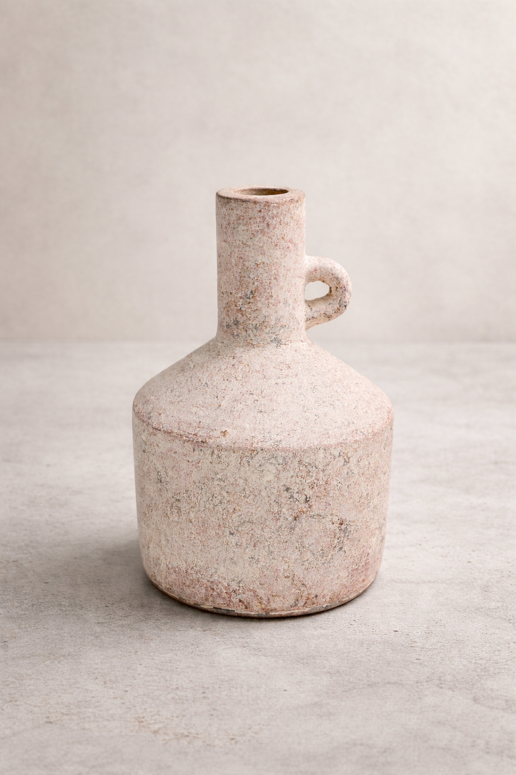 A handmade, sandy-beige terracotta vase with a rounded body and narrow neck, standing alone on a smooth concrete floor against a soft linen-colored wall in a minimalist studio setting.