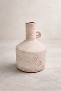 A handmade, sandy-beige terracotta vase with a rounded body and narrow neck, standing alone on a smooth concrete floor against a soft linen-colored wall in a minimalist studio setting.