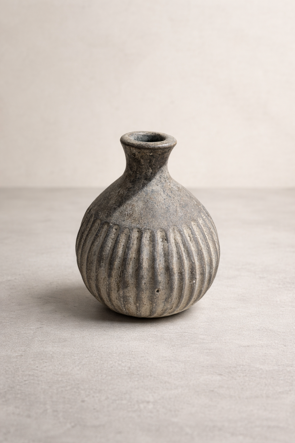 A textured gray ceramic vase with vertical ridges sits centered on a smooth light concrete floor against a neutral linen-colored wall in a softly lit studio setting.