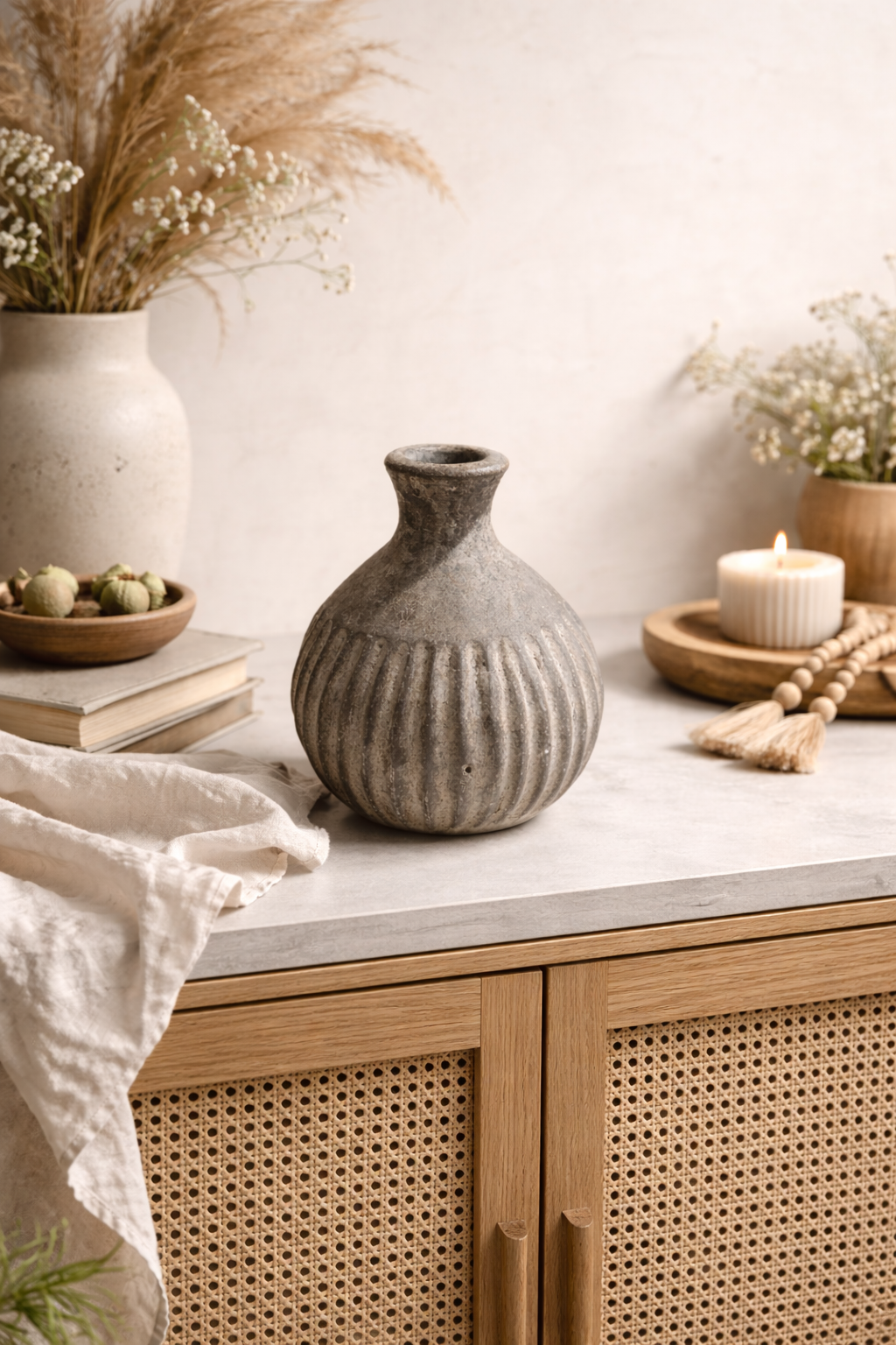 A ribbed gray ceramic vase sits on a smooth light concrete surface, surrounded by soft linen fabric, a lit candle on a wooden tray, small bowls, and dried grasses against a warm linen-colored wall, creating a calm, natural, and cosy studio scene.