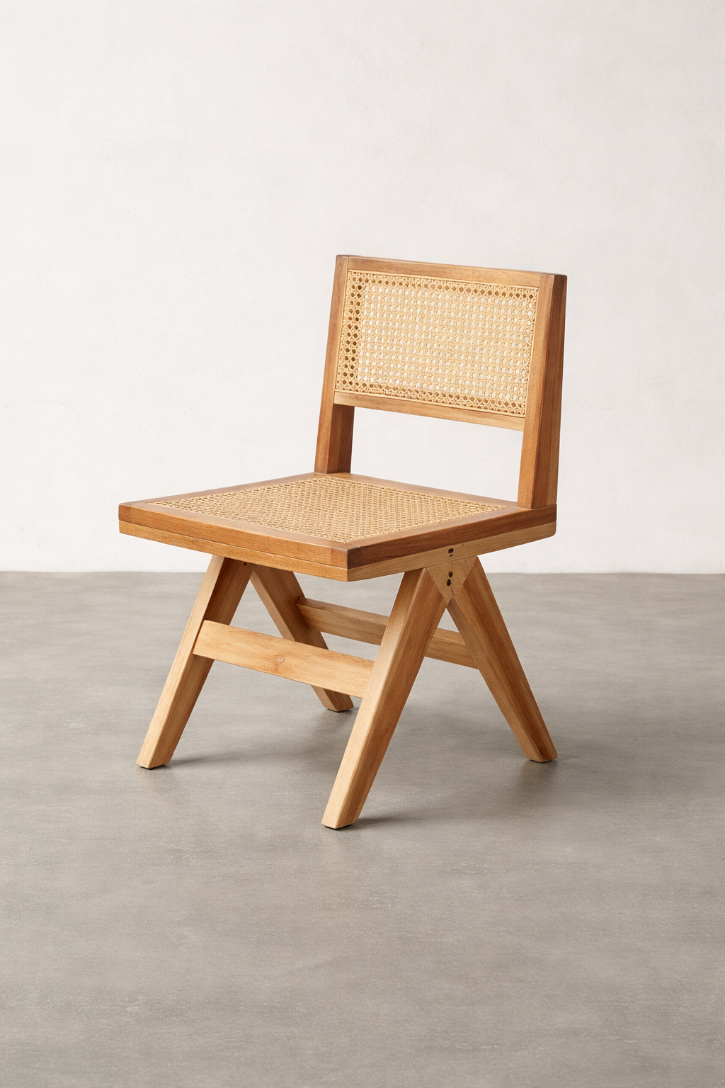 A teak dining chair with a woven cane seat and back is shown alone in a studio setting, resting on a smooth concrete floor against a soft linen-colored wall under gentle, even light.