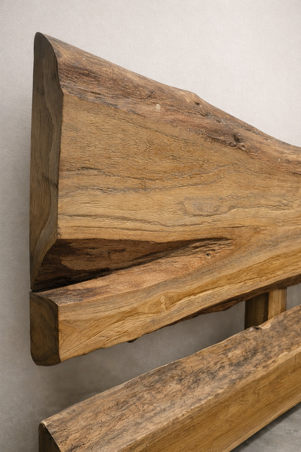 Close-up of a handcrafted live-edge wooden headboard showing rich natural grain and organic texture, photographed in a minimalist studio with a soft linen-colored wall and smooth concrete floor.