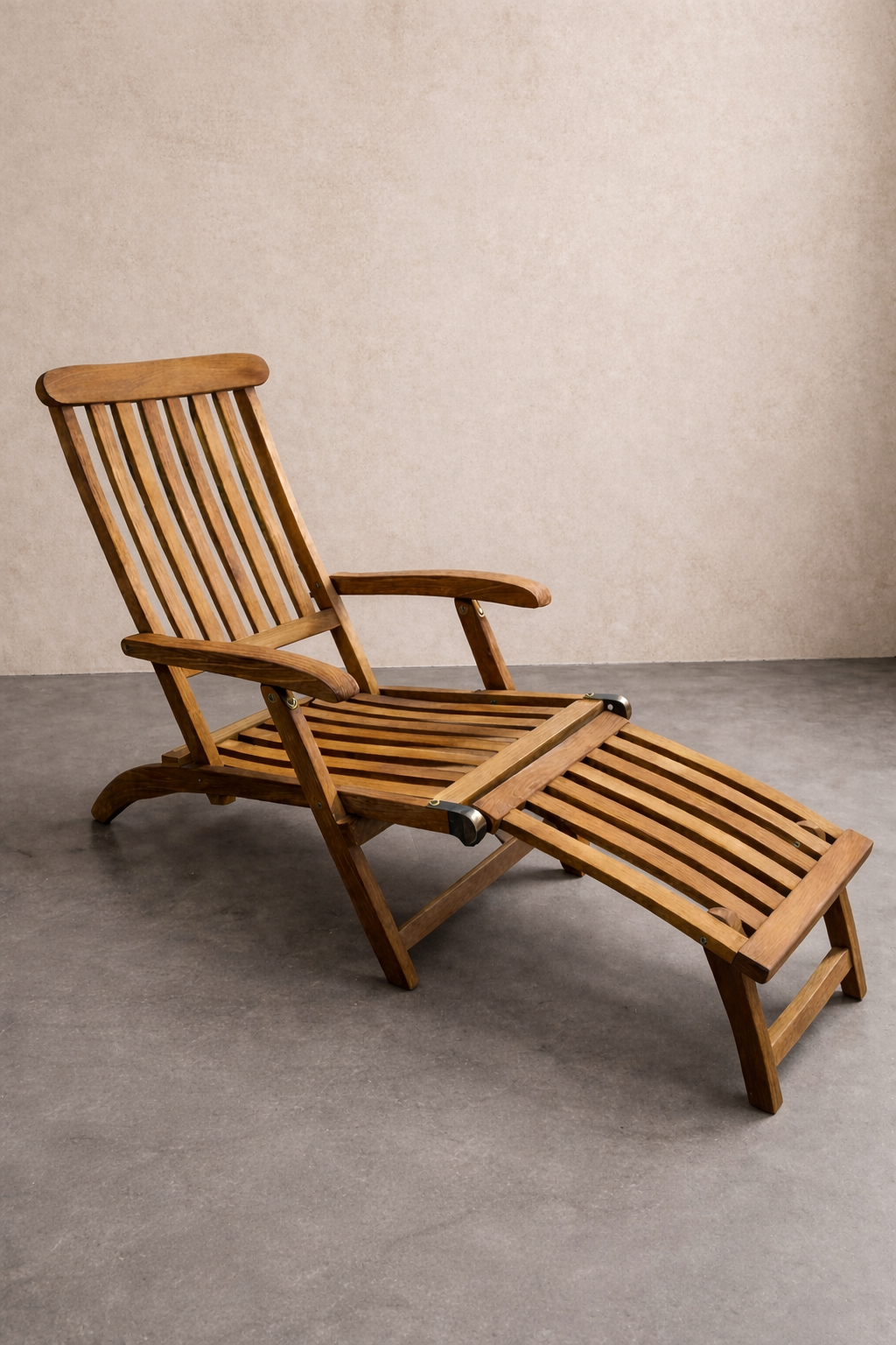 A teak reclining lounge chair with slatted seat and footrest sits on a smooth grey concrete floor against a textured linen-colored wall, softly lit to highlight the wood grain in a calm, minimalist studio setting.