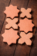 Six handmade terracotta tiles—star and cross shapes—are arranged in two columns on a dark wooden surface, showing warm earthy tones and natural texture.