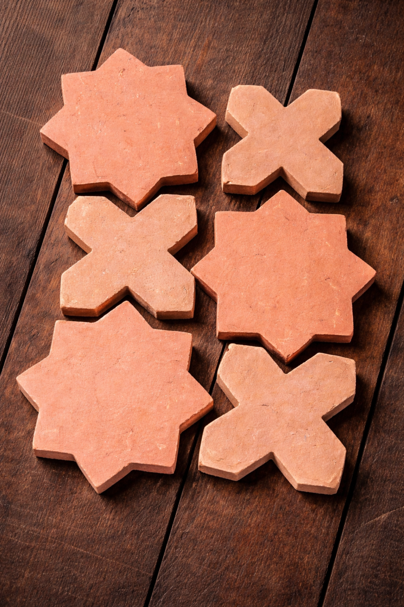 Six handmade terracotta tiles—star and cross shapes—are arranged in two columns on a dark wooden surface, showing warm earthy tones and natural texture.